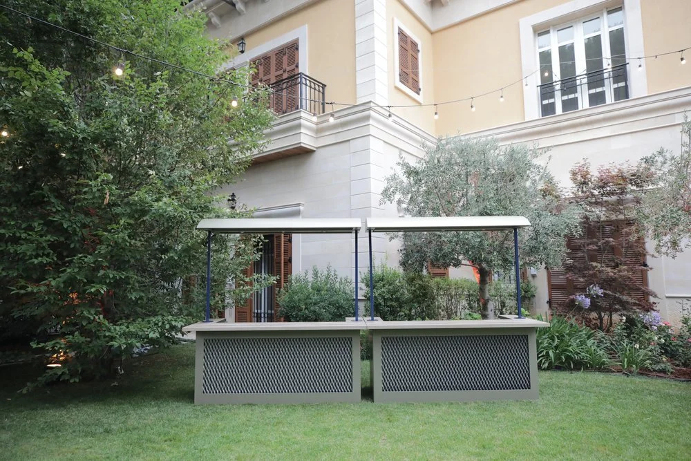 Outdoor scene with a white mobile food stand or cart on a grassy lawn, surrounded by trees and plants, with a beige building in the background and string lights overhead.