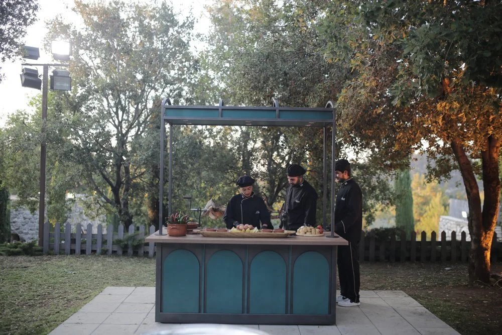 Three chefs standing behind a blue outdoor food cart preparing food outdoors in a park with trees and a fence.