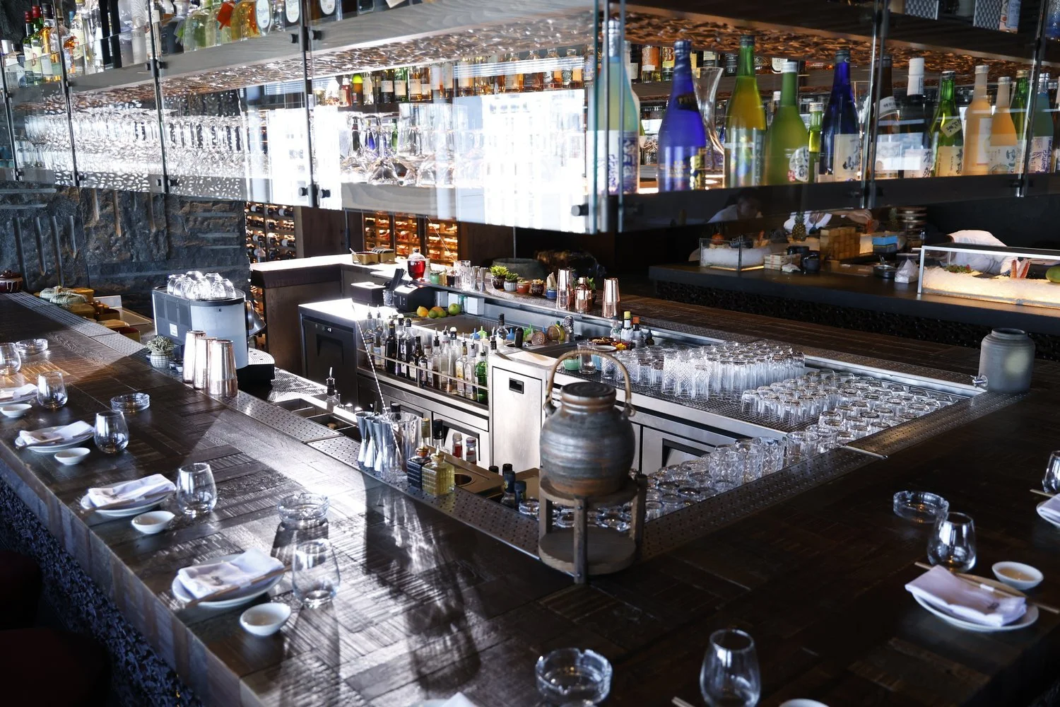 Upscale restaurant bar with a wooden counter set for dining, glassware, napkins, and dishes, featuring a well-stocked bar with bottles, glasses, and bar equipment, illuminated by natural light.