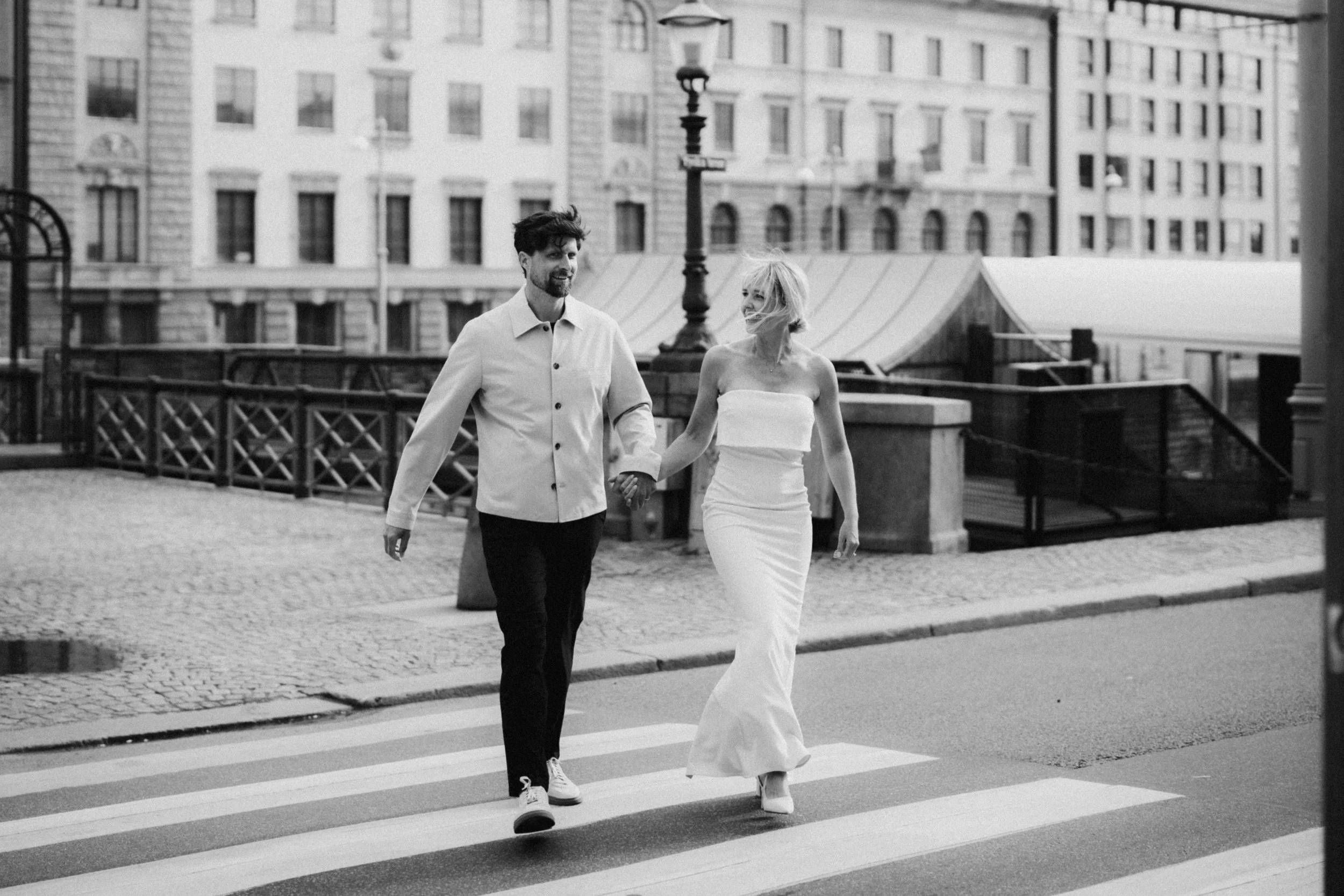 En svartvit bild av ett par, troligen brud och brudgum, som går över ett övergångsställe i en stadsmiljö med gamla byggnader i bakgrunden.