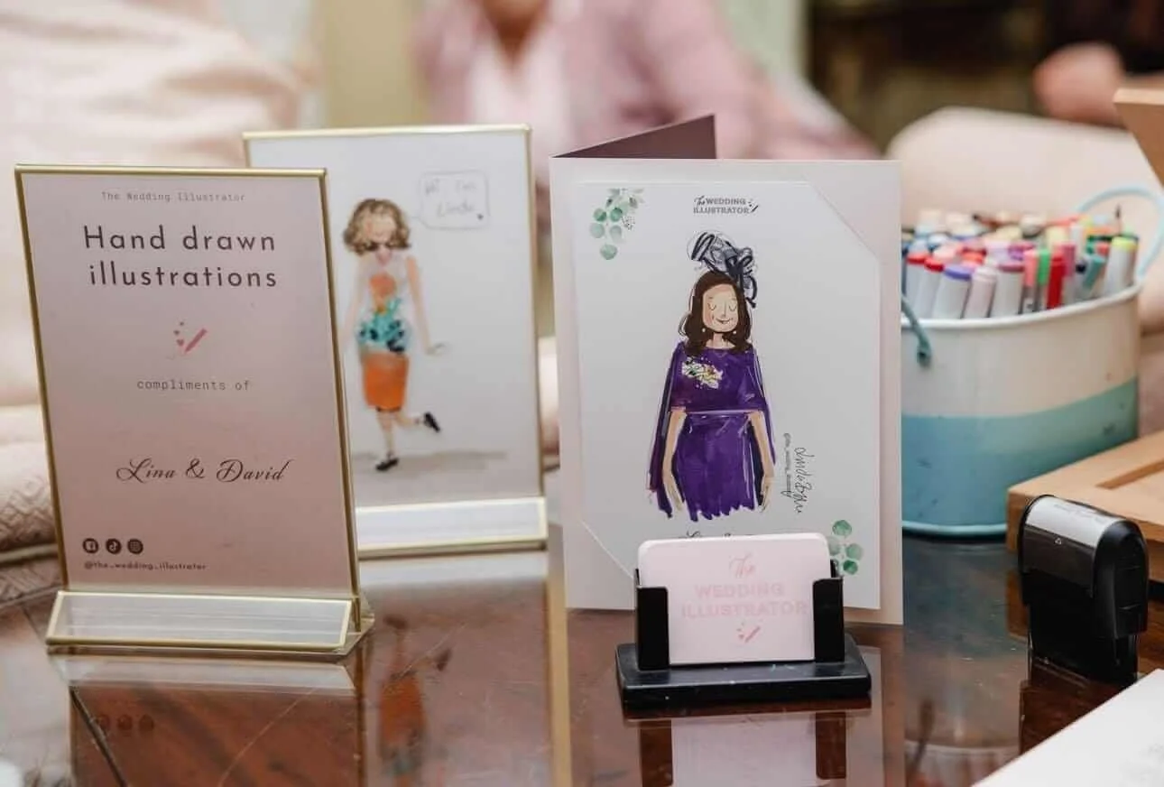 Display of handwritten wedding illustrations and cards on a table, with markers and a stamp nearby.
