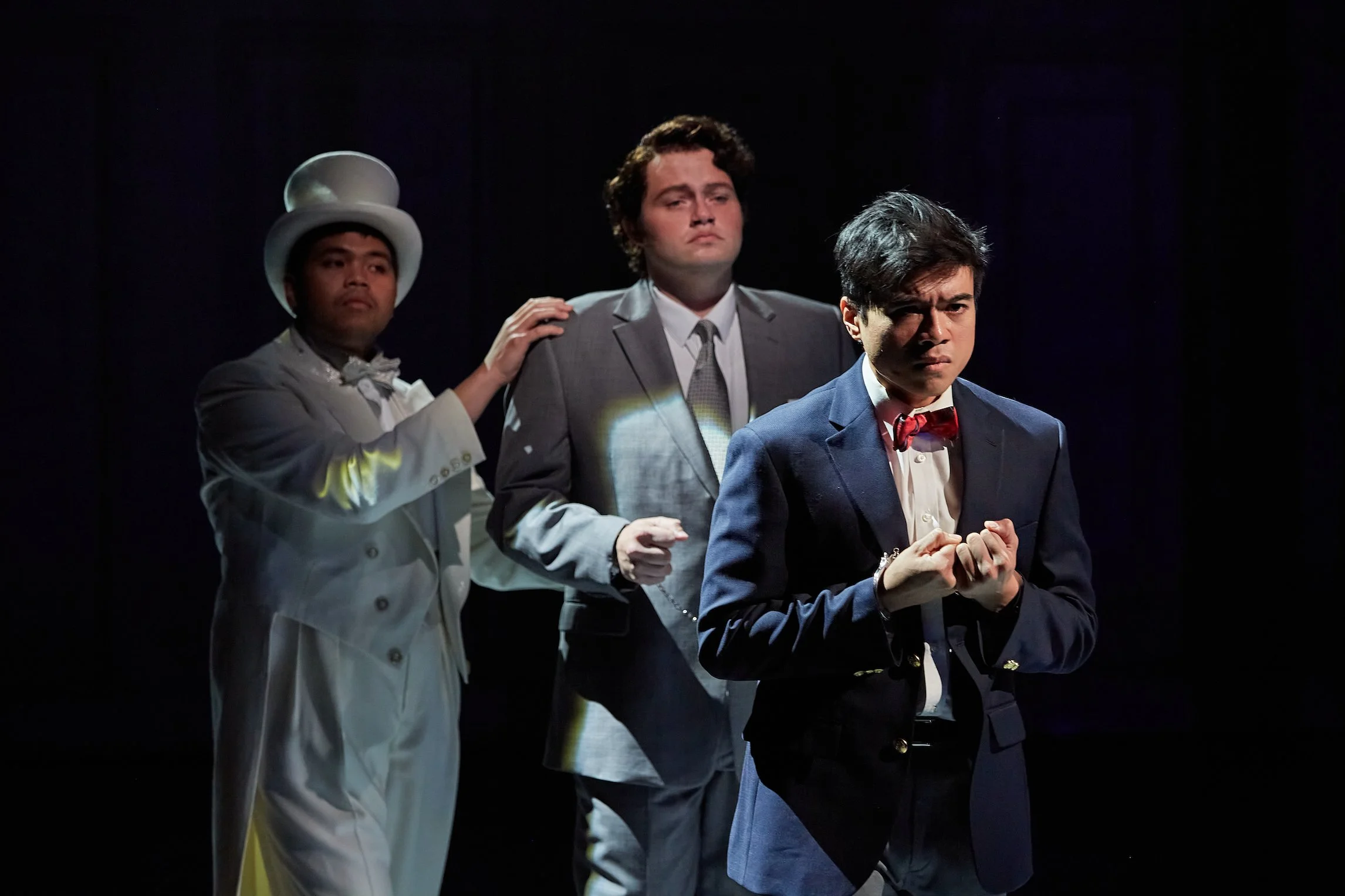 Three men in formal attire are staged in a dramatic scene. The man in the foreground wears a dark blazer and red bow tie, with clenched fists and a serious expression. Behind him, a man in a gray suit looks solemn, and another in a white suit with a