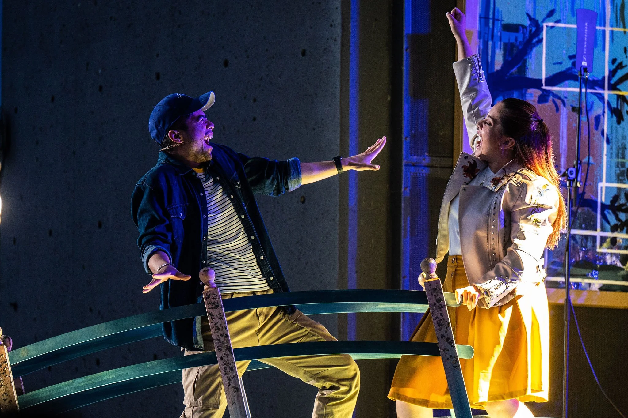 Two people smiling and playfully reaching towards each other on stage, one wearing a baseball cap and striped shirt, the other in a beige coat and yellow skirt, with colorful stage lighting and background setup.