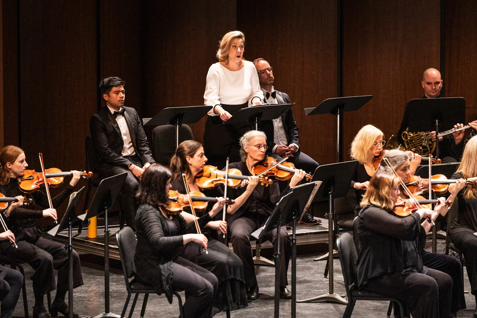Orchestra performing on stage with a woman conductor standing at her podium, surrounded by musicians playing violins and other instruments, in a concert hall with wooden walls.