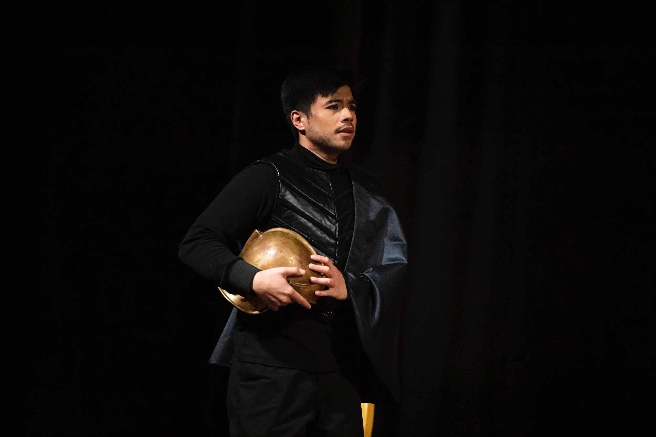 A man dressed in black holding a brass helmet, standing against a dark background.