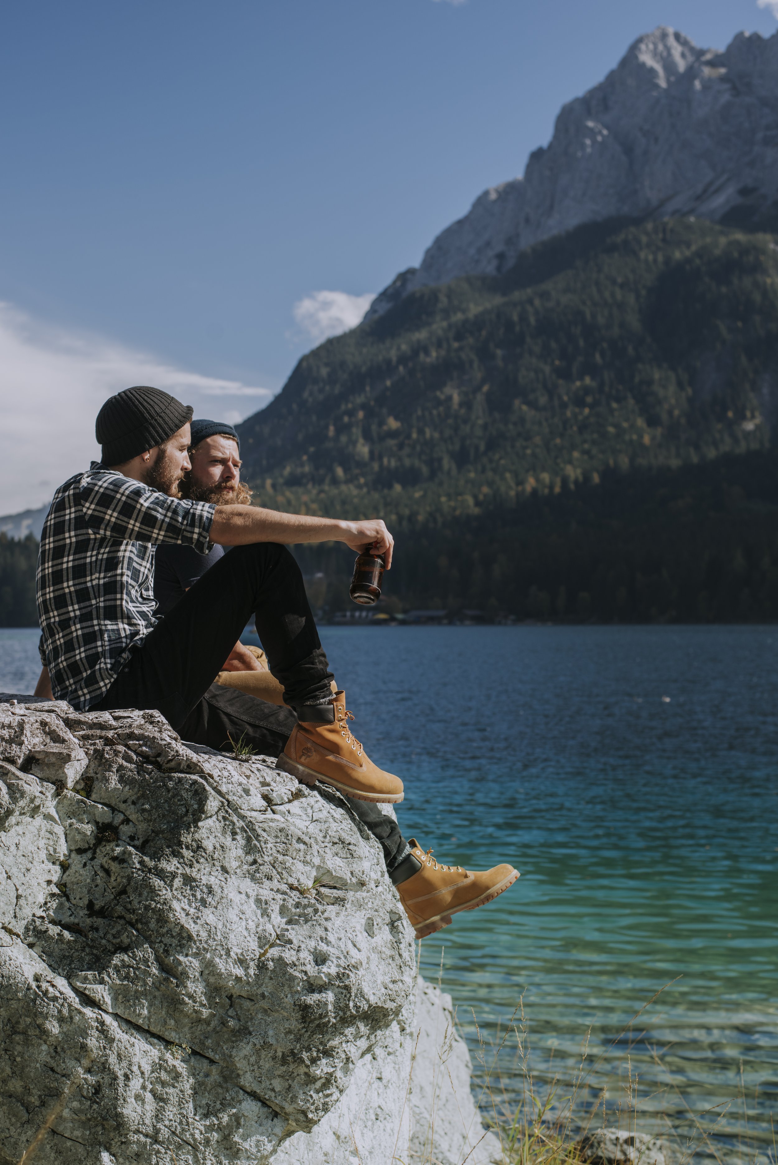 Zwei Männer sitzen auf einem Felsen am See mit Bergen im Hintergrund, einer hält eine Bierflasche, beide tragen Beanies und Wanderstiefel.