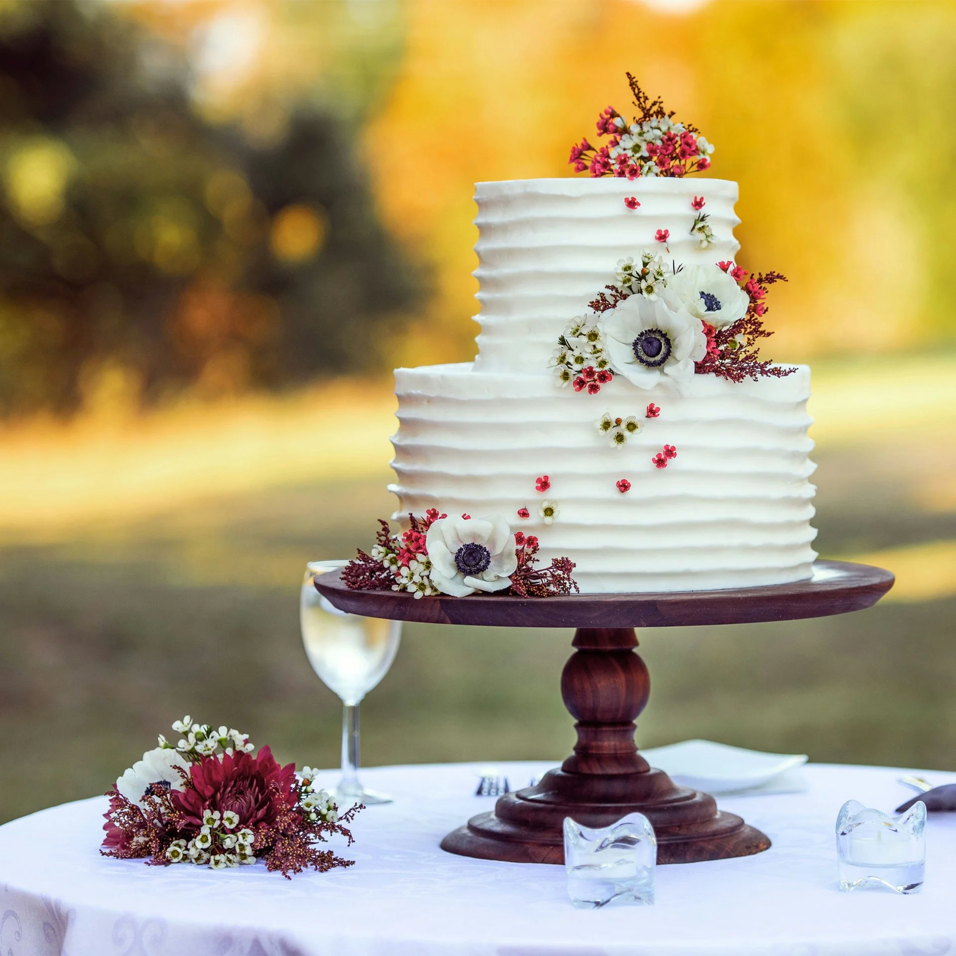 Two-tiered white wedding cake decorated with fresh flowers and placed on a wooden cake stand, outdoors with blurred autumn trees in the background.