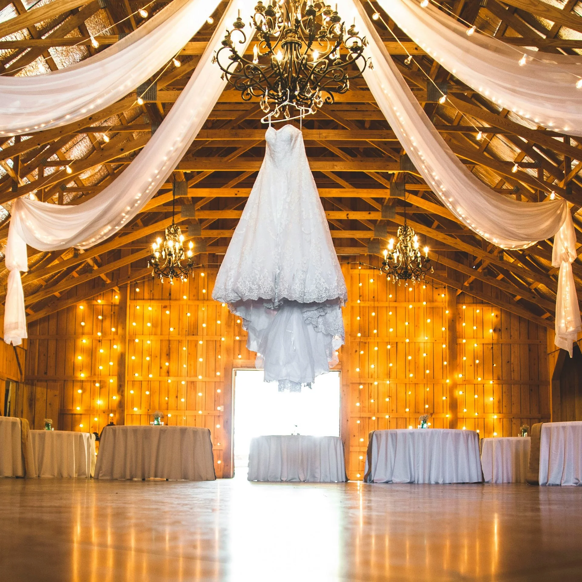 Wedding dress hanging from a chandelier in a rustic barn decorated with string lights and draped fabric