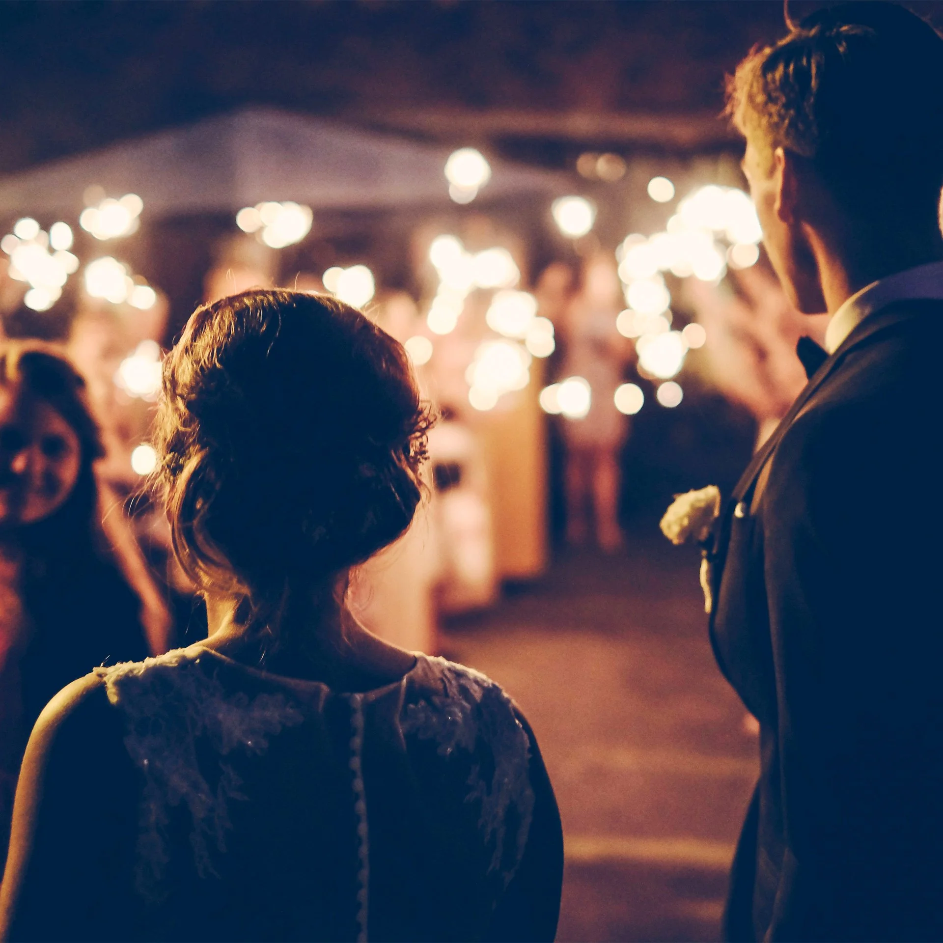 A bride and groom at their wedding reception surrounded by guests, with the bride facing away and the groom looking at her, illuminated by warm string lights.