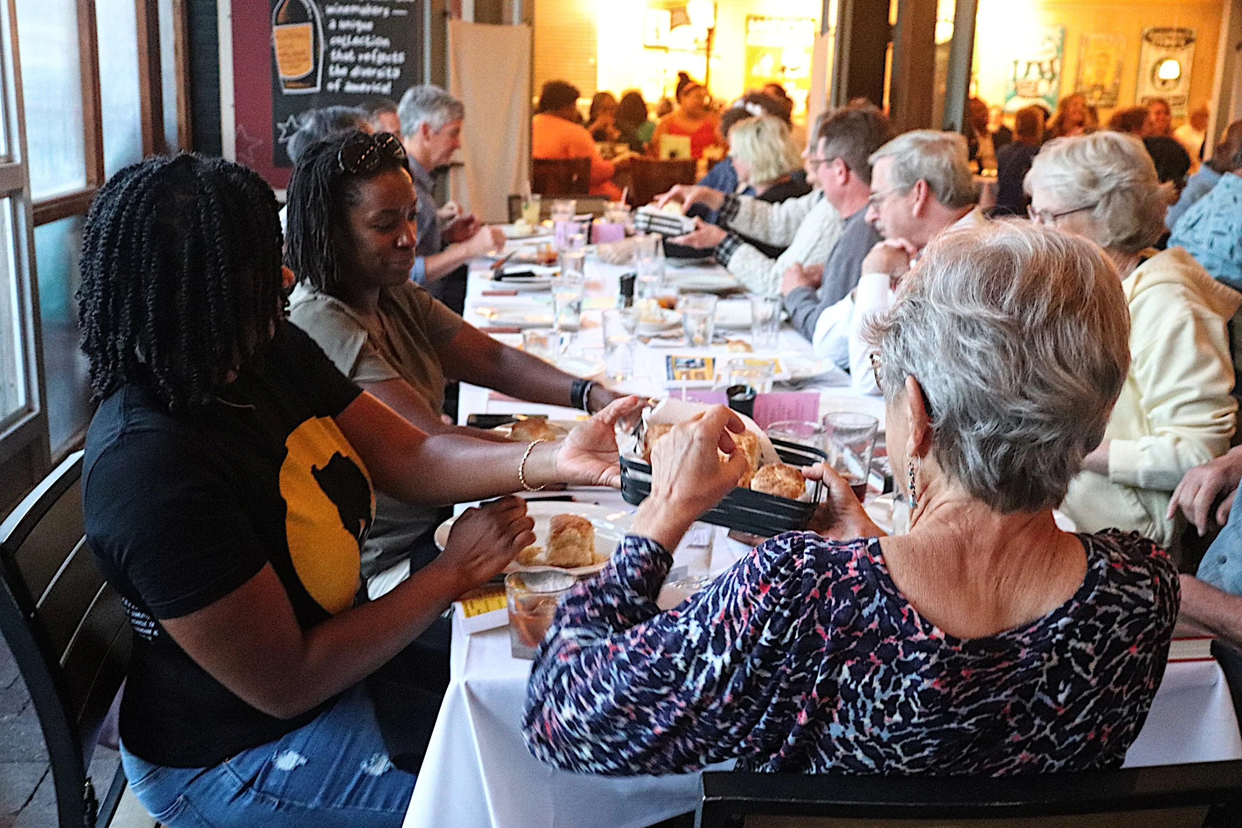 A diverse group of people dining together at a long restaurant table, sharing food and engaging in conversation.
