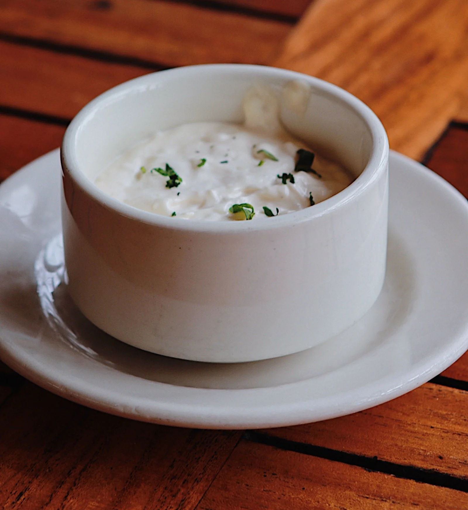 A white cup of creamy tartar sauce with chopped herbs served on a white plate on a wooden table.