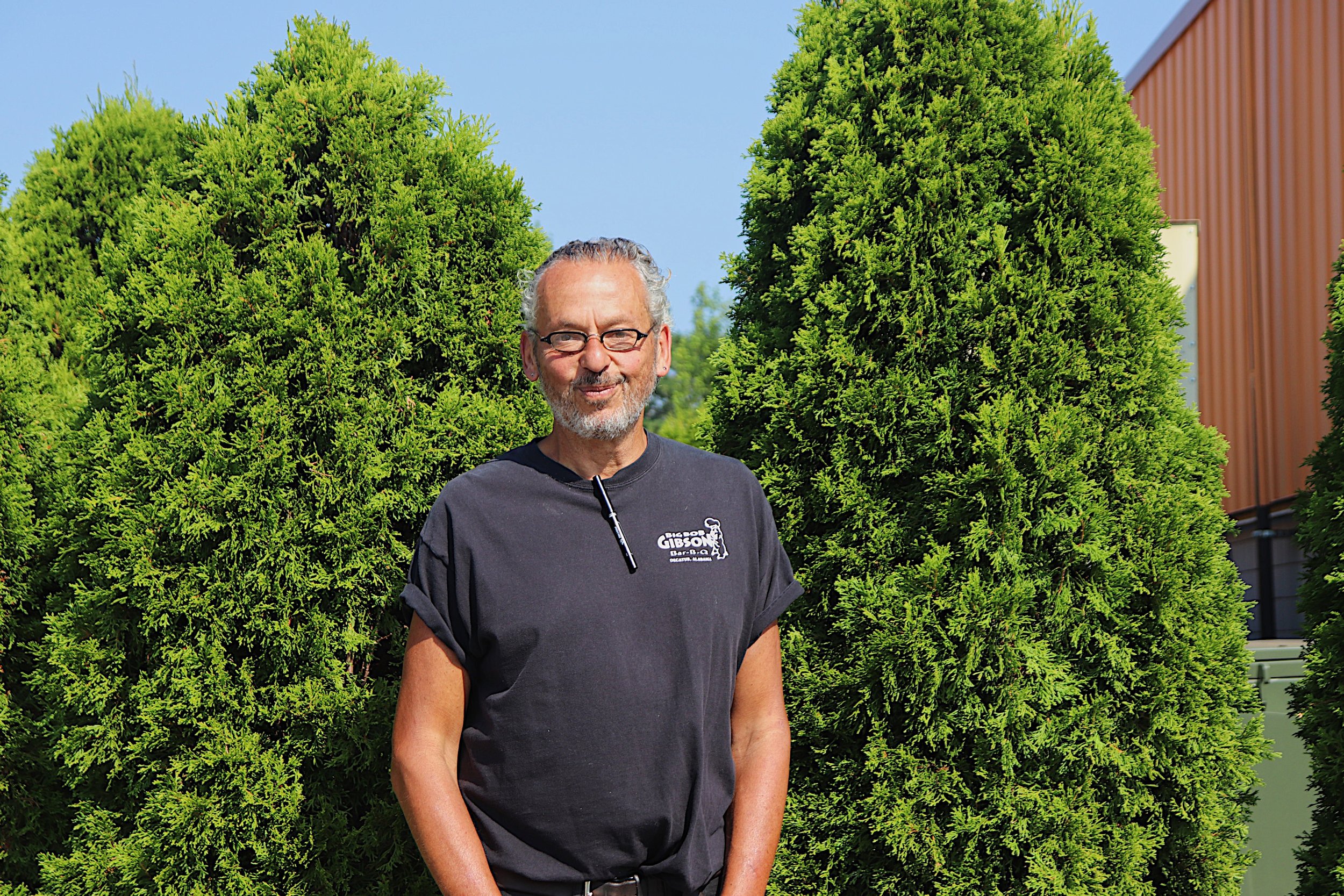 A man with glasses and gray hair standing outdoors near green bushes, wearing a black T-shirt with white text and a logo.