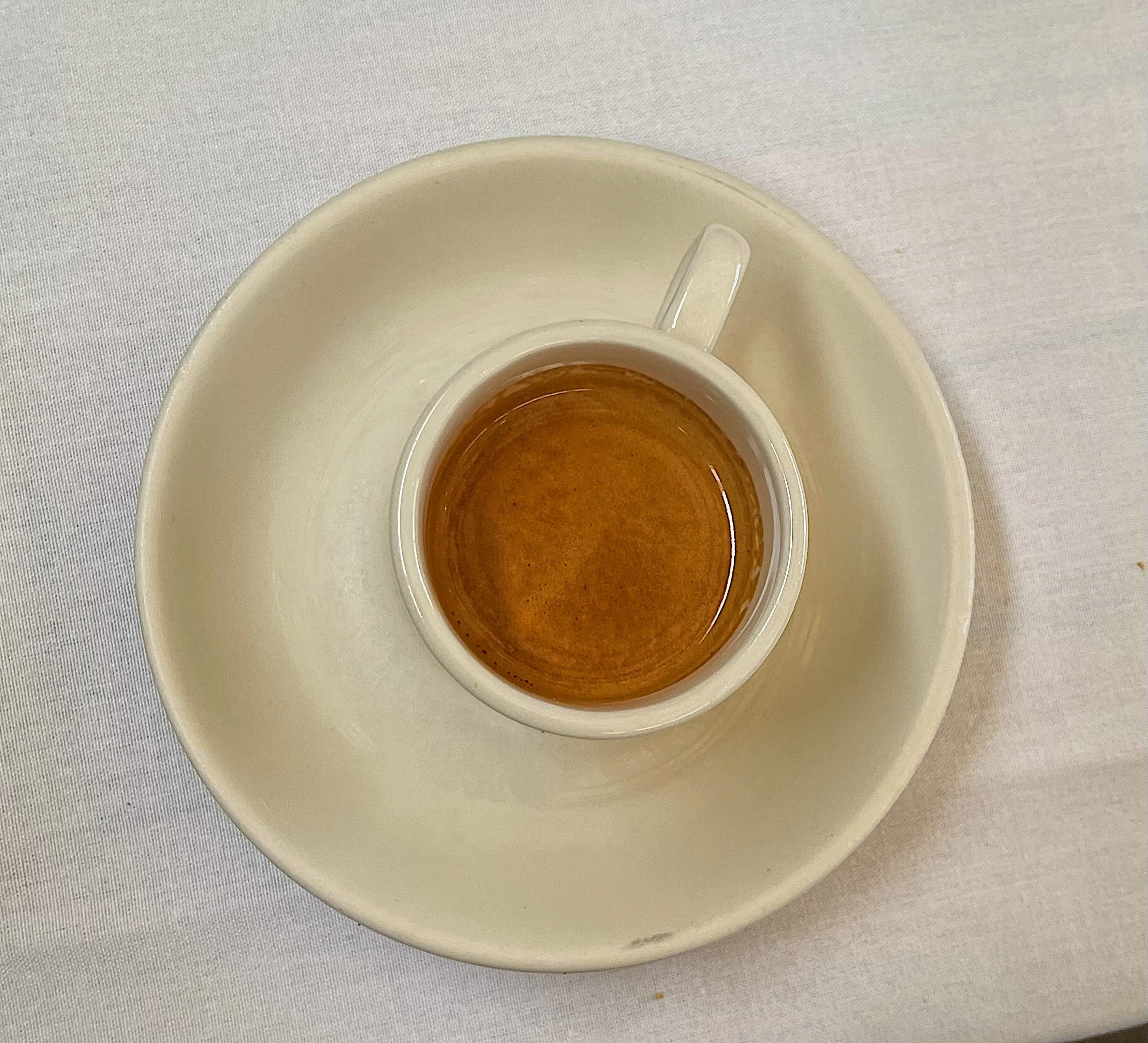 A small white espresso cup filled with dark coffee on a matching white saucer placed on a light-colored tablecloth.