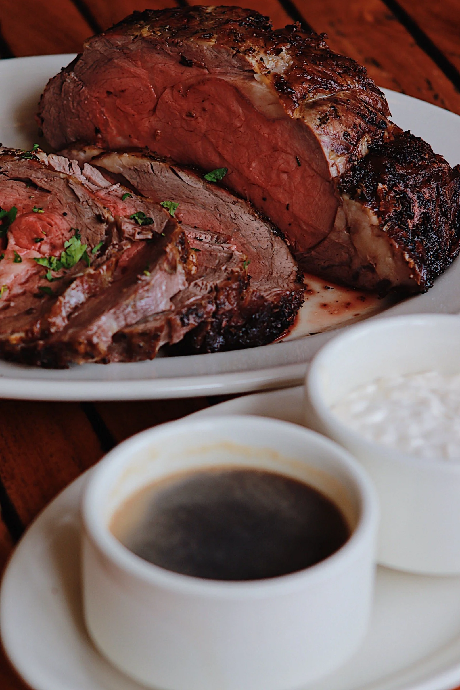 Cooked prime rib roast with slices on a white plate, served with side bowls of black coffee and horseradish sauce.