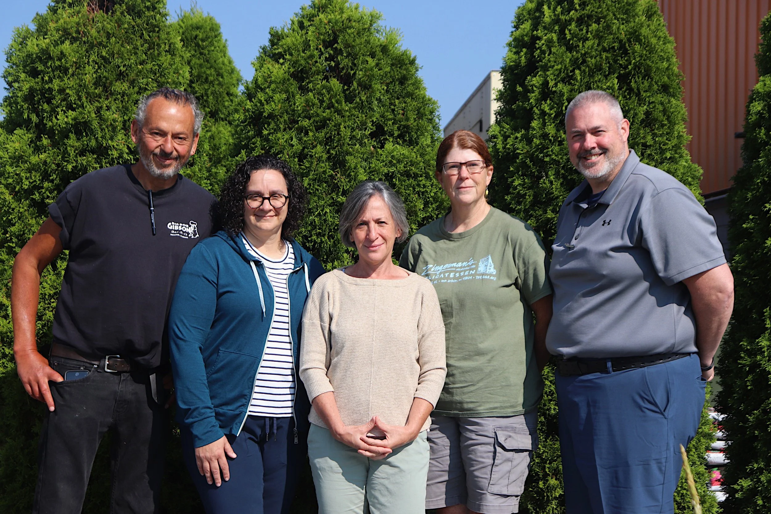 Group of five people standing outdoors in front of green trees, smiling at the camera.