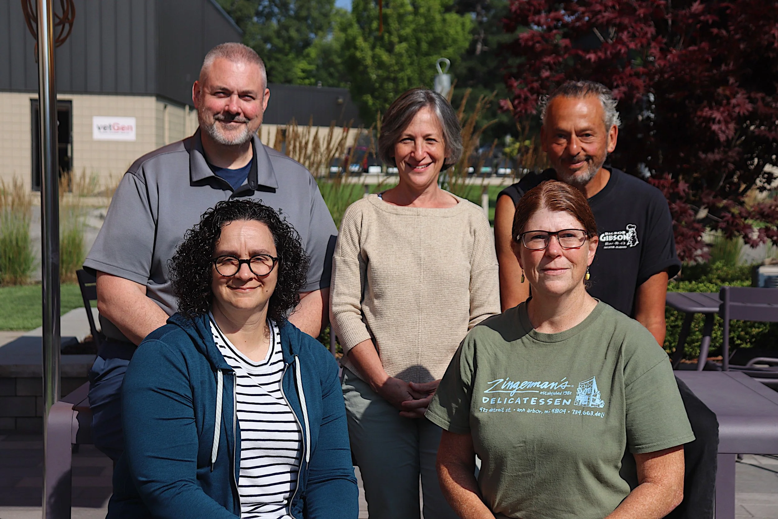 Six people posing outdoors in a garden setting, standing and sitting in two rows. The group includes four women and two men, all smiling at the camera, with trees and buildings in the background.