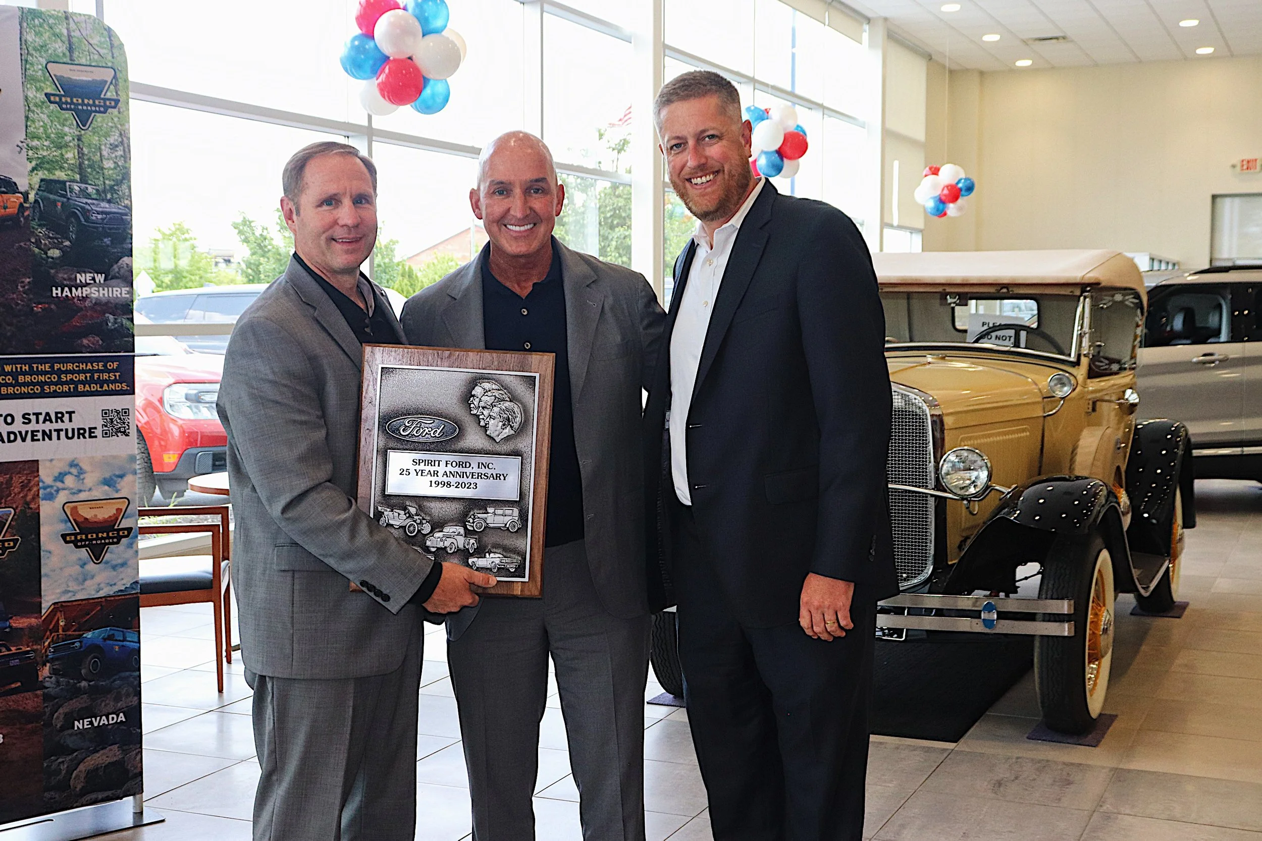 Three men in suits at a car dealership celebrating 25 years of Spirit Ford, Inc., holding a framed plaque, with a vintage yellow Ford Model T car and promotional banners in the background.