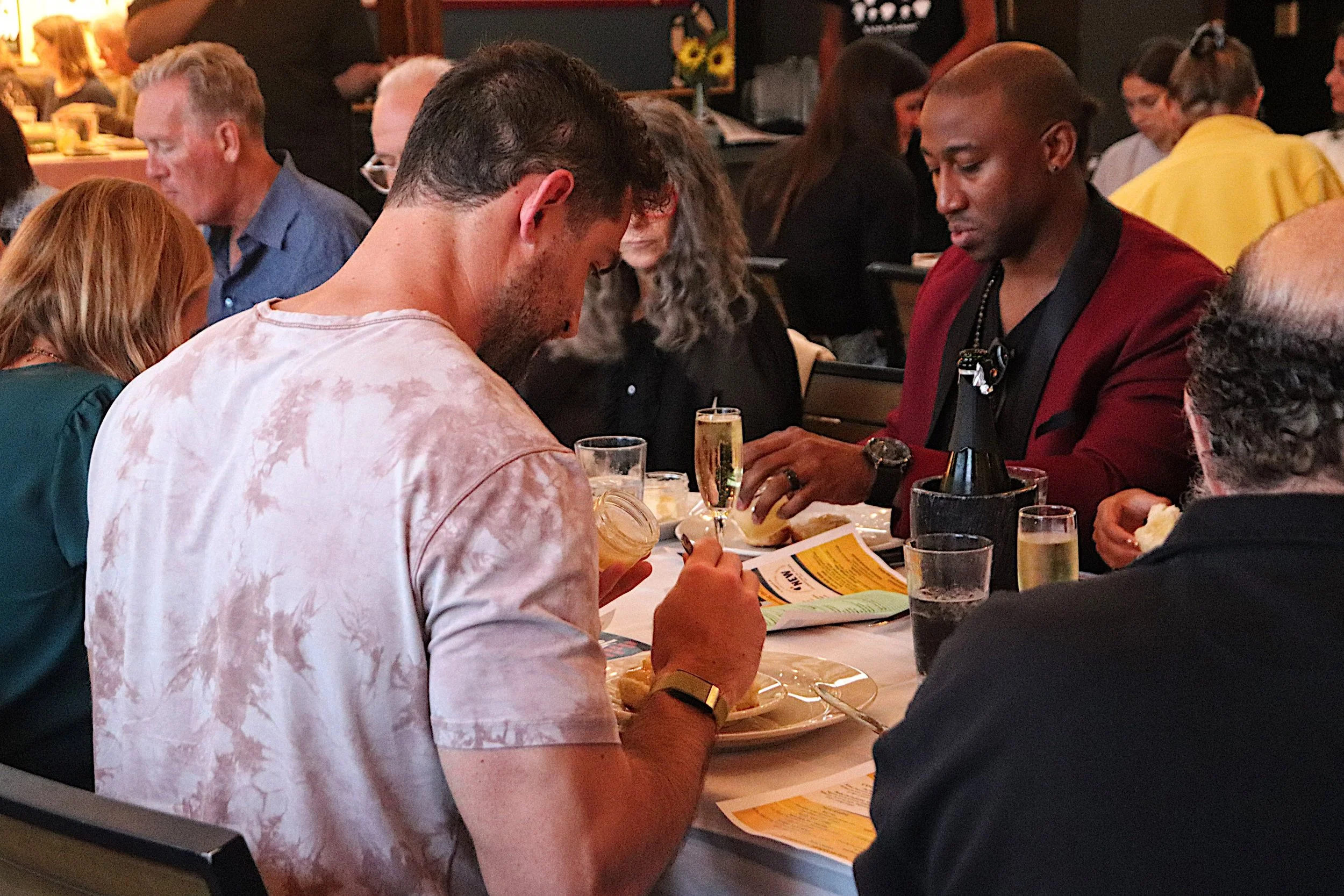 People gathered around a table at a restaurant, eating and drinking, with various drinks including champagne, wine, and water visible.