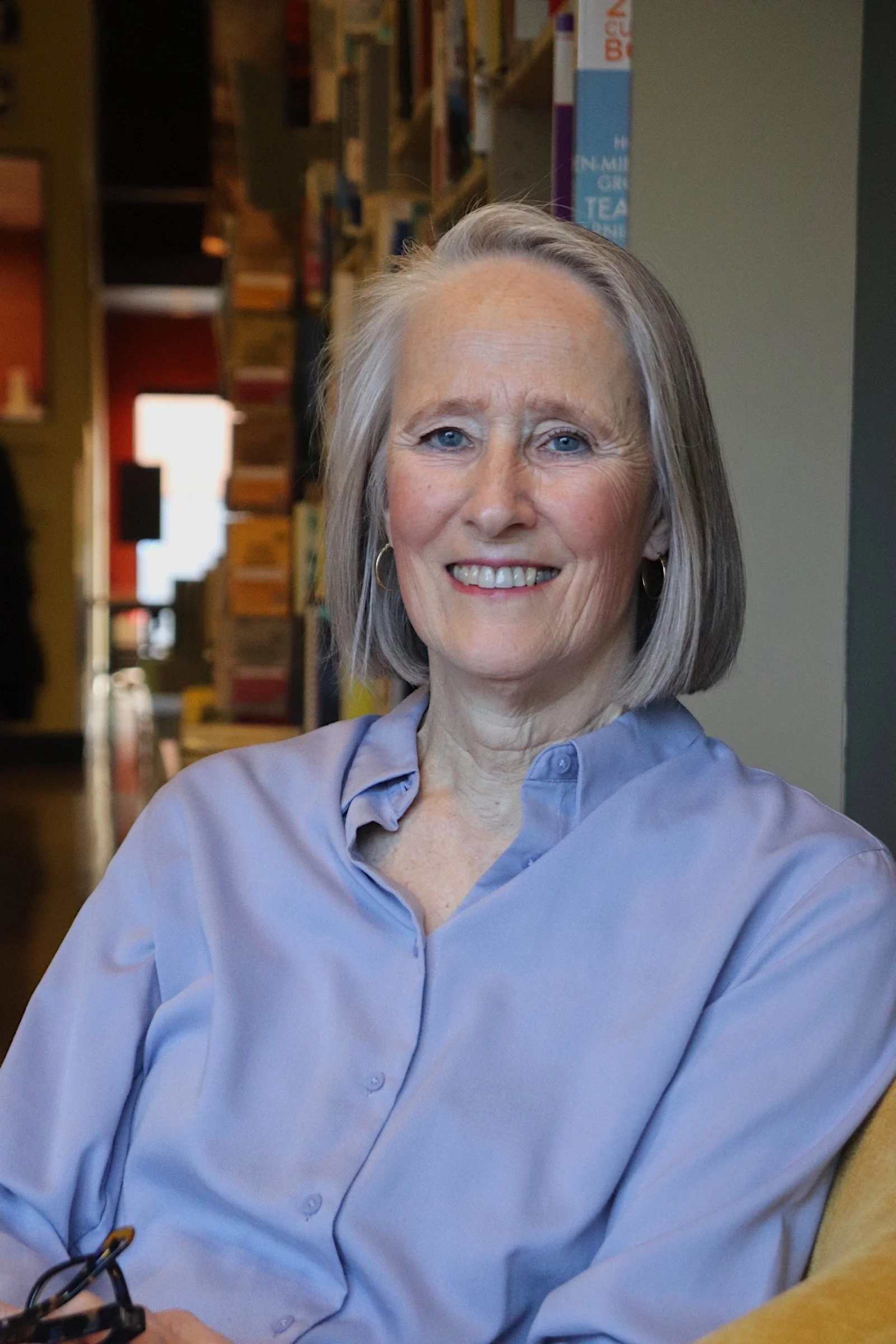 A smiling older woman with gray hair and blue eyes, wearing a light blue button-up shirt, sitting indoors with bookshelves in the background.