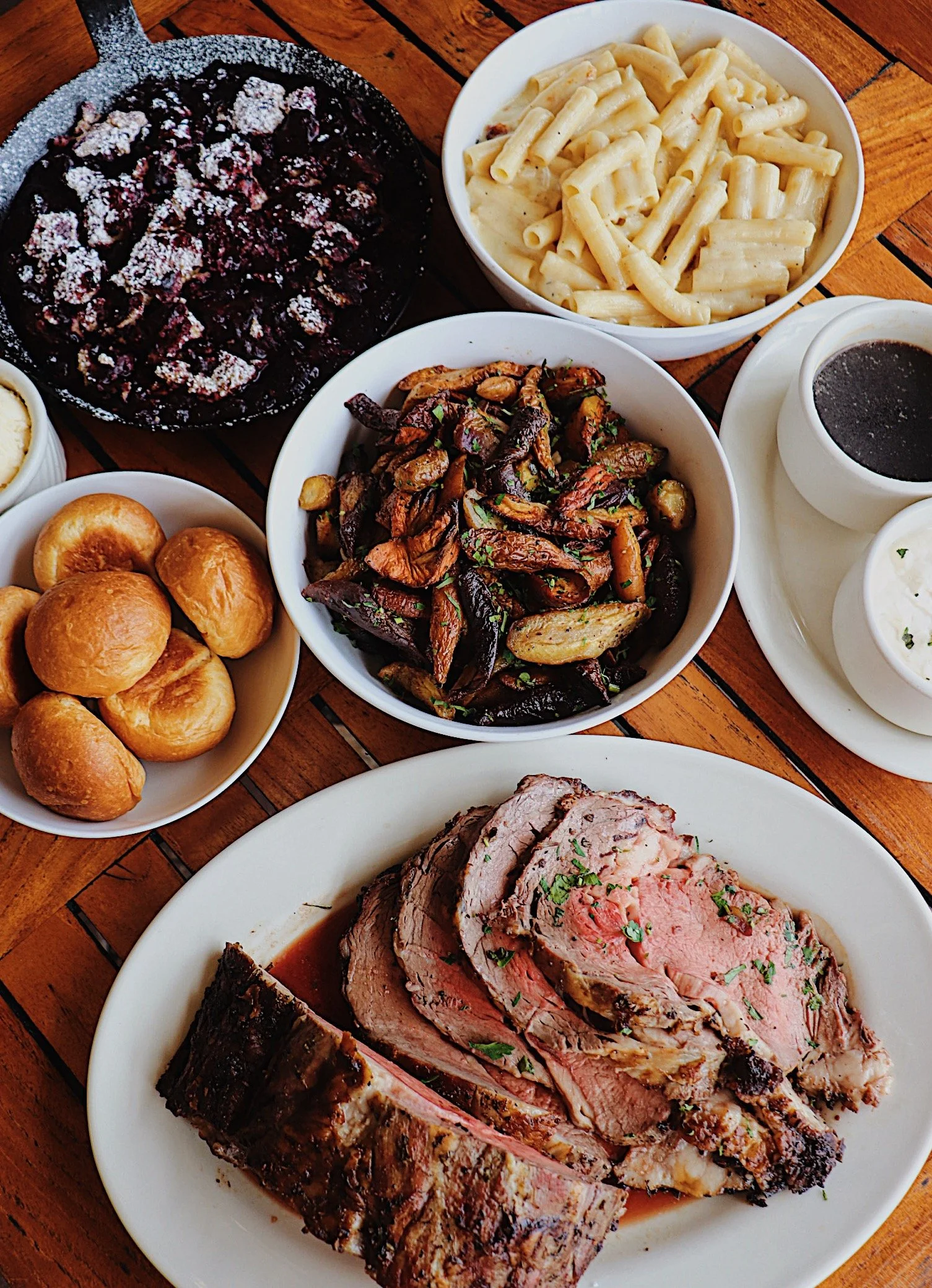 A spread of dishes including roasted beef, pasta, roasted vegetables, bread rolls, cherry pie, and condiments on a wooden table.