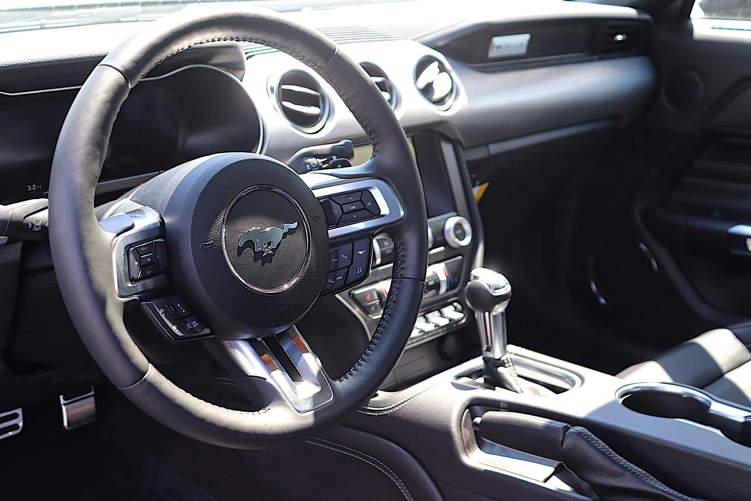 Interior of a Ford Mustang car showing a black steering wheel with a Mustang logo, dashboard with air vents, gear shift, and control buttons.