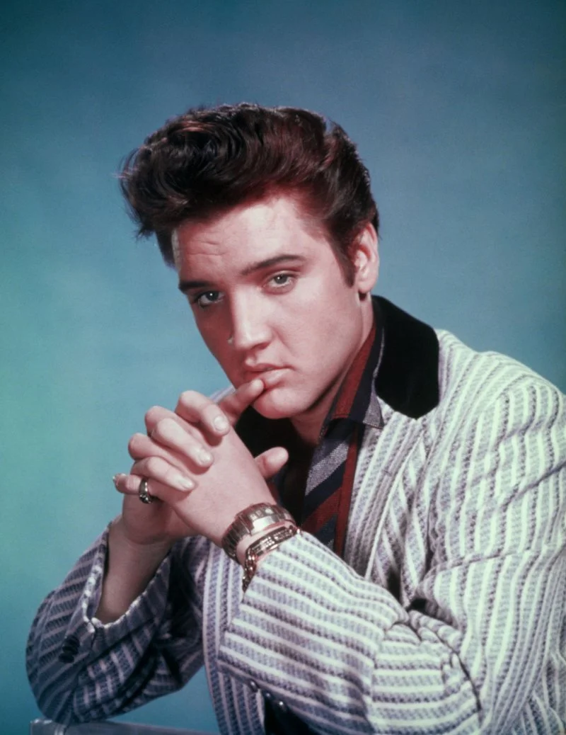 A young man with styled brown hair wearing a striped blazer, a patterned shirt, and a red and navy striped tie, with his hands clasped near his face, looking directly at the camera against a plain blue background.