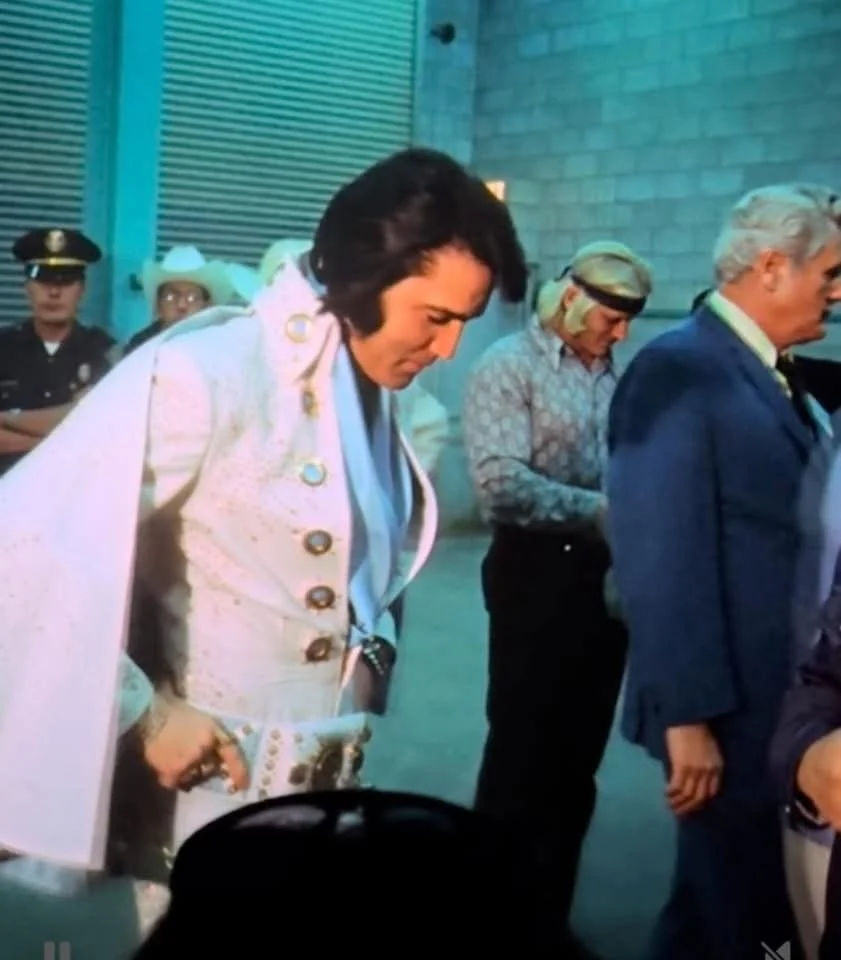 Group of people standing in line, including a man dressed as Elvis Presley and police officer in uniform in background.
