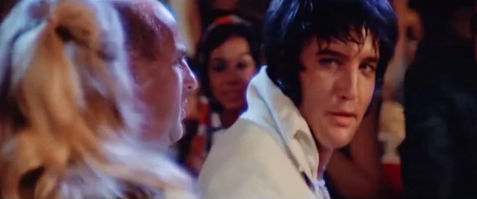 A scene from a movie with a man and a woman in a social setting, possibly a bar or club, with a woman with red hair talking to a woman with long blonde hair, and a man with dark hair turned toward the camera.