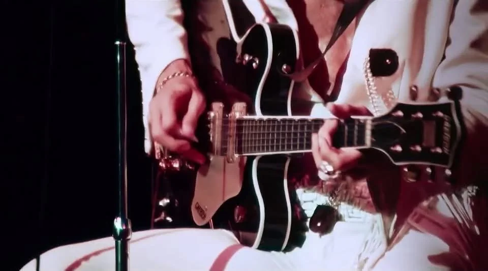 Person playing an acoustic guitar with a white body and black edges, wearing a white outfit with pearl and metallic accents.