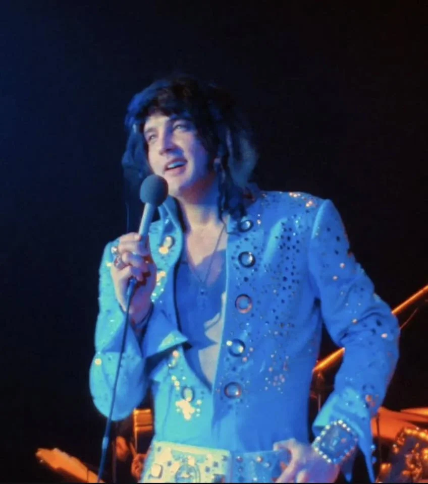 Person dressed as Elvis Presley performing on stage with a microphone, wearing a blue sequin outfit and a wig.