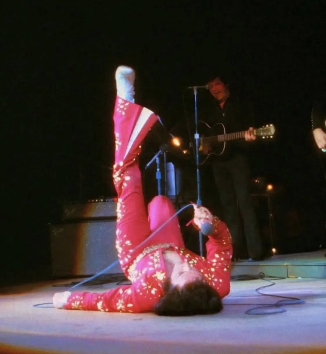 A person lying on the floor in a red holiday sweater with white snowflake patterns, holding a microphone, in front of a performer playing an acoustic guitar and singing into a microphone on stage.