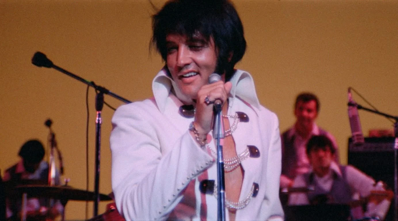 Elvis Presley performing on stage in a white jumpsuit, holding a microphone, with backup musicians in the background.