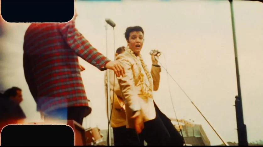 A young man singing or speaking into a microphone on an outdoor stage with a surprised or expressive look, surrounded by others.