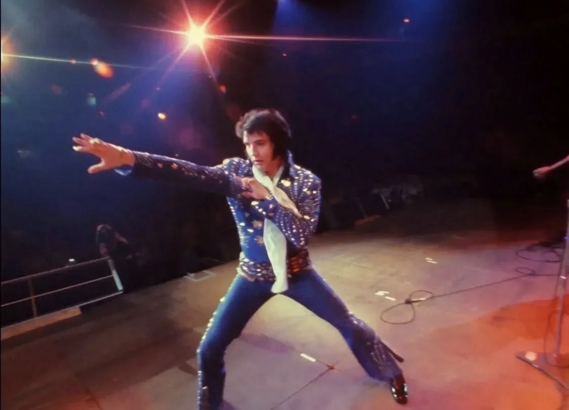 Elvis Presley performing on stage in a sparkly blue jacket, blue jeans, and a white scarf, striking a classic pose with one arm extended and the other near his chest.