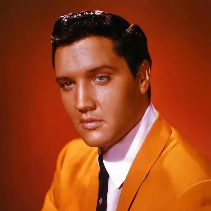 Portrait of a young man with slicked-back hair wearing a yellow suit and a white shirt against a red background.