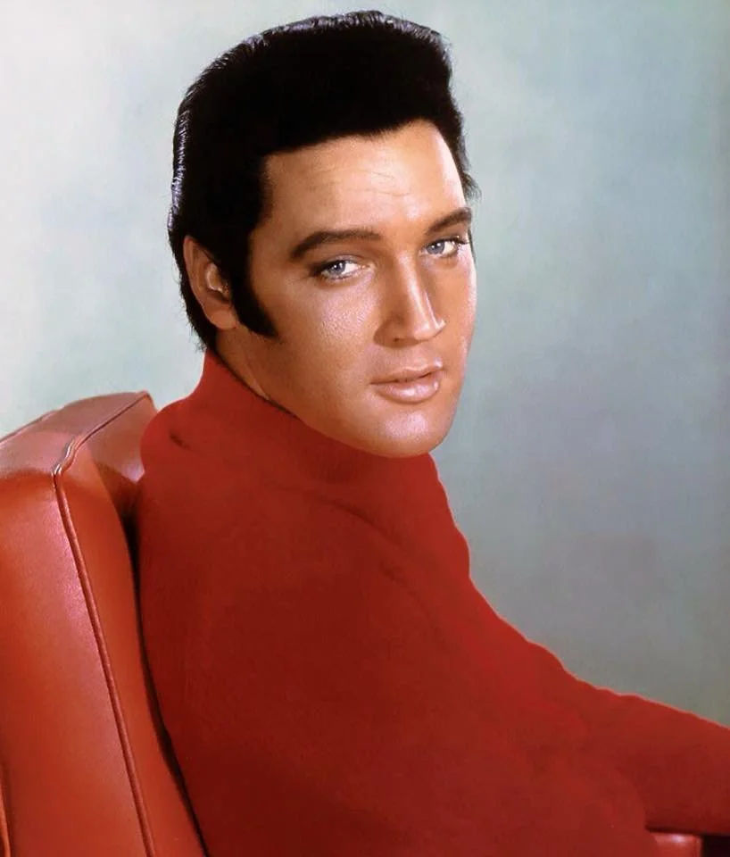 Portrait of a man with dark hair wearing a red turtleneck sitting in a red chair, looking at the camera with a slight smile.