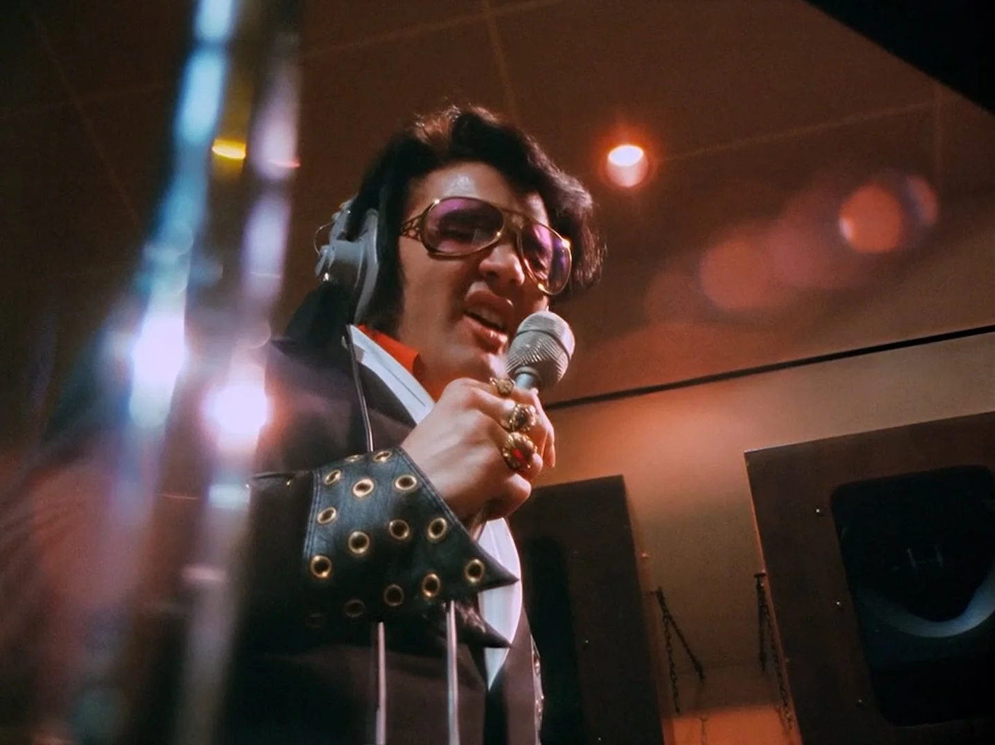 A person dressed as Elvis Presley, wearing large sunglasses and a black leather jacket with metal eyelets, singing into a microphone while wearing headphones in a dimly lit room.