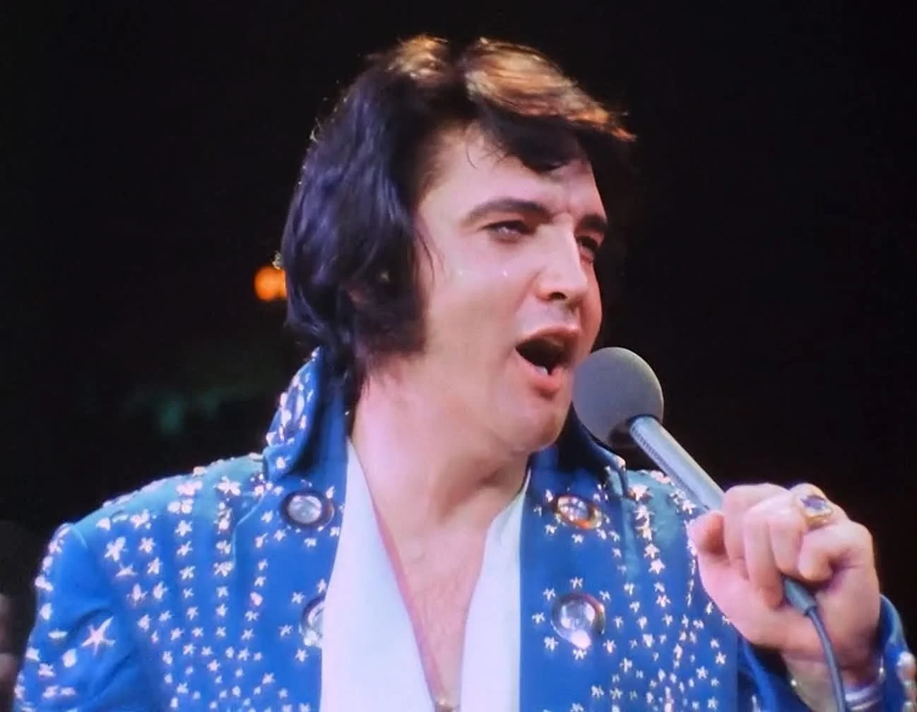 A man dressed as Elvis Presley singing into a microphone while on stage.