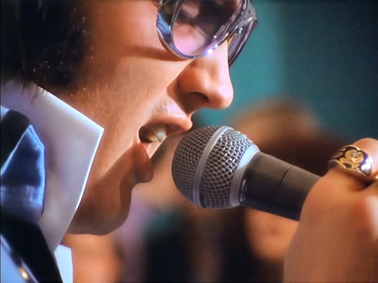 Close-up of a woman singing or speaking into a microphone, wearing glasses and a black jacket with a white shirt underneath.