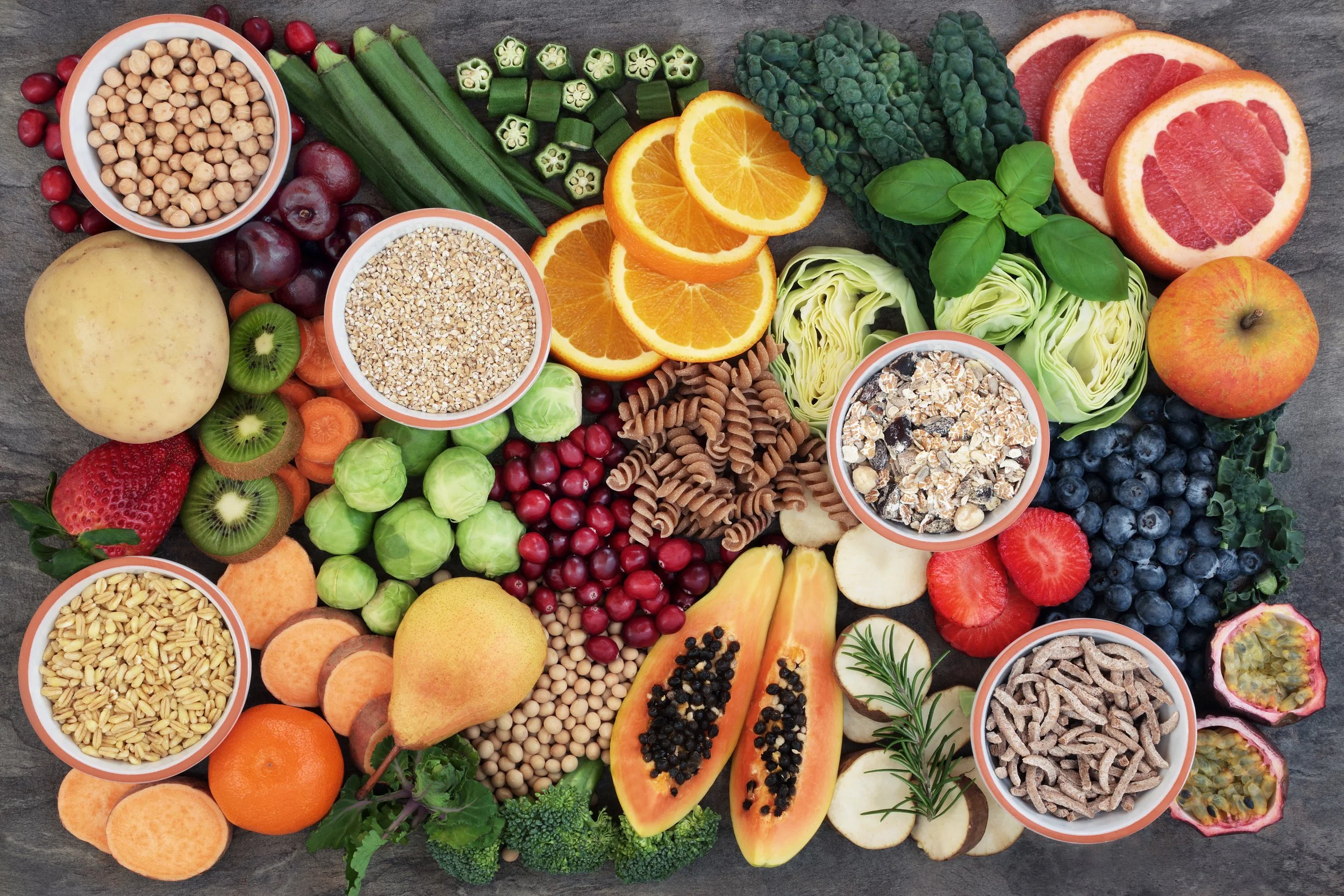 Assorted fresh fruits, vegetables, grains, and nuts showcased on a wooden surface, including oranges, grapefruit, papaya, blueberries, strawberries, kiwi, apple, Brussels sprouts, zucchini, okra, cherry tomatoes, and various grains and nuts in bowls.