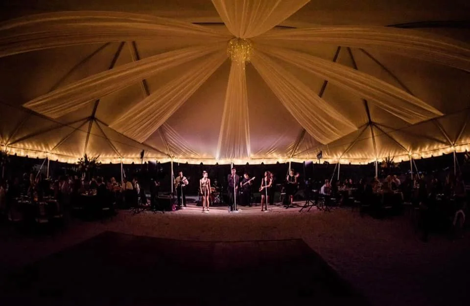 A band performing under a large, elegantly draped tent at night with ambient lighting and tables around the perimeter.