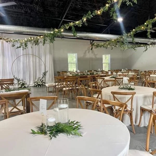 Wedding reception hall with round tables, wooden chairs, white tablecloths, greenery decorations, hanging lights, and a circular backdrop.