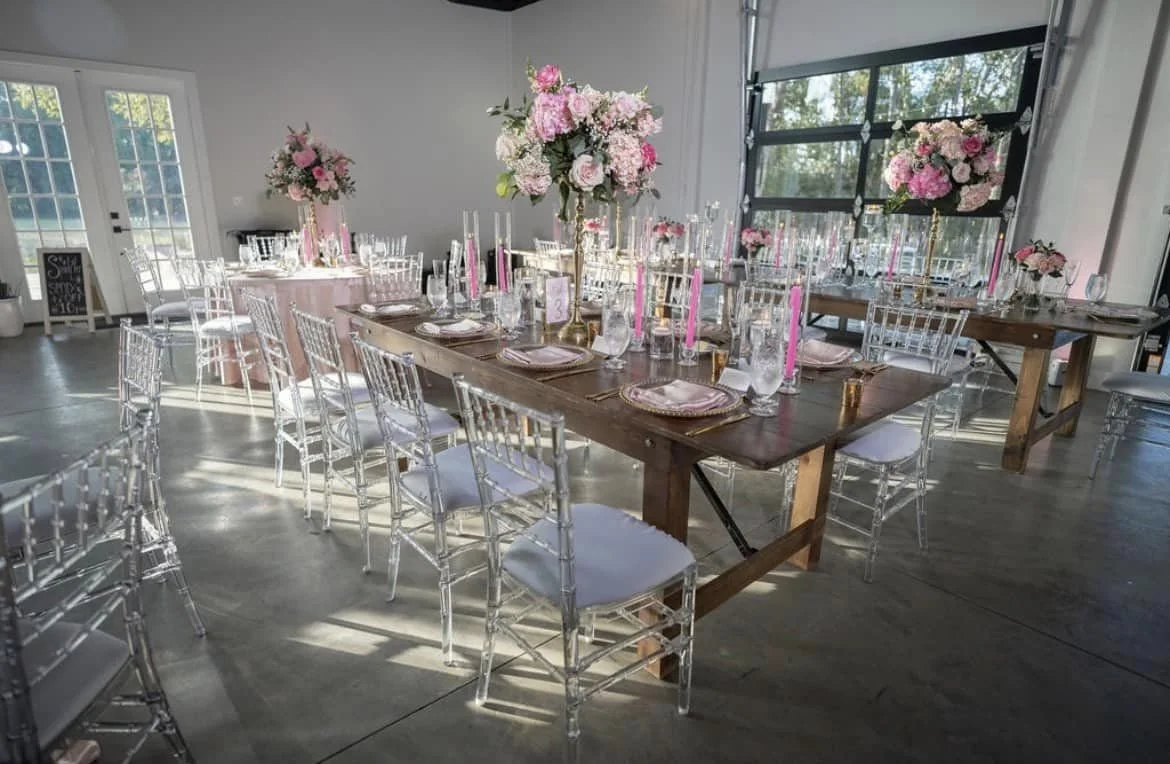 Elegant indoor event setup with long wooden tables, clear chairs, and pink floral arrangements.