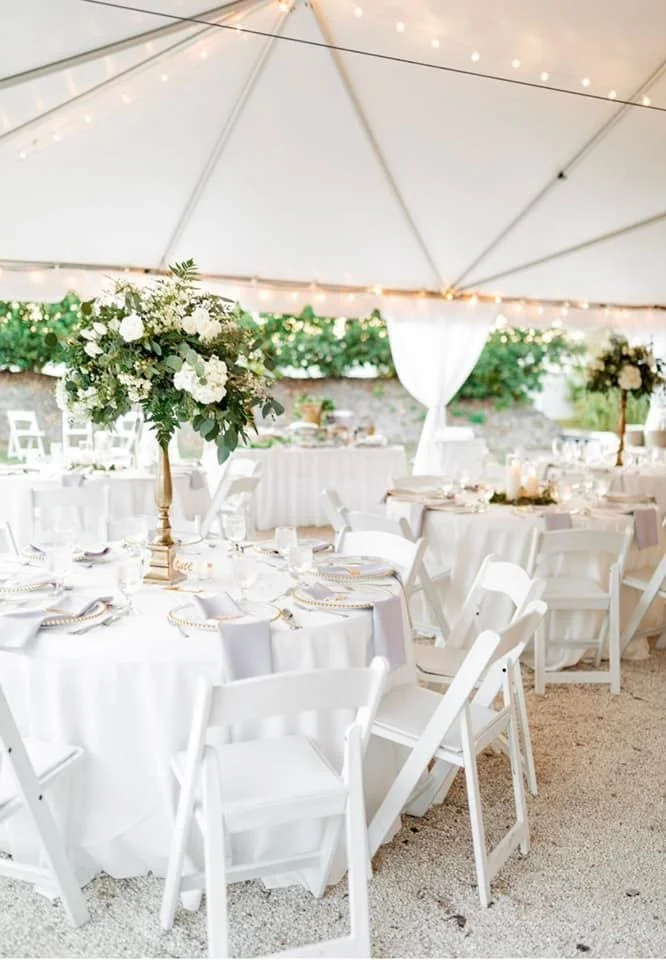 Elegant outdoor wedding reception setup under a tent with white round tables, white chairs, lush floral centerpieces, and string lights.