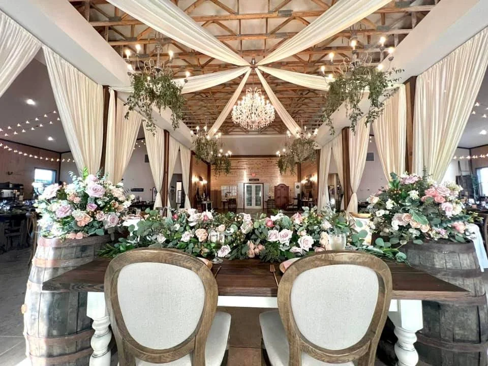 Elegant wedding reception setup with wooden chairs, floral centerpieces, draped fabric ceiling, chandeliers, and decorative greenery.