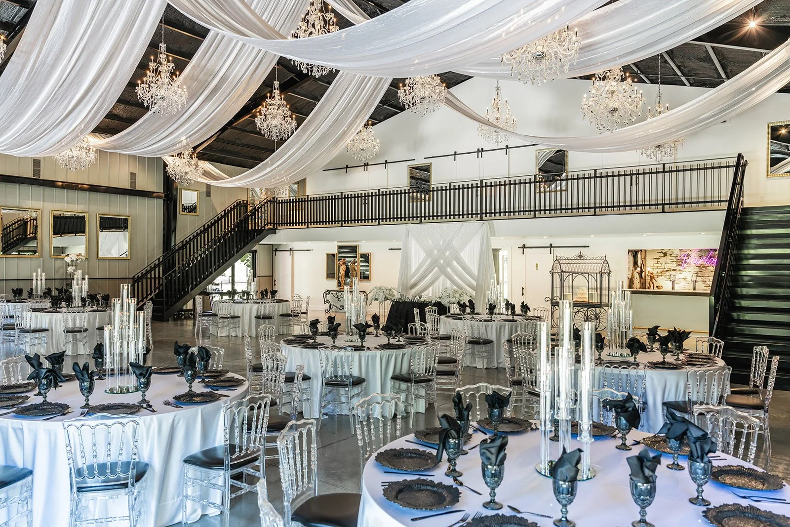 Elegant wedding venue setup with round tables decorated in black and white, translucent chairs, chandeliers, and draped fabric on the ceiling.
