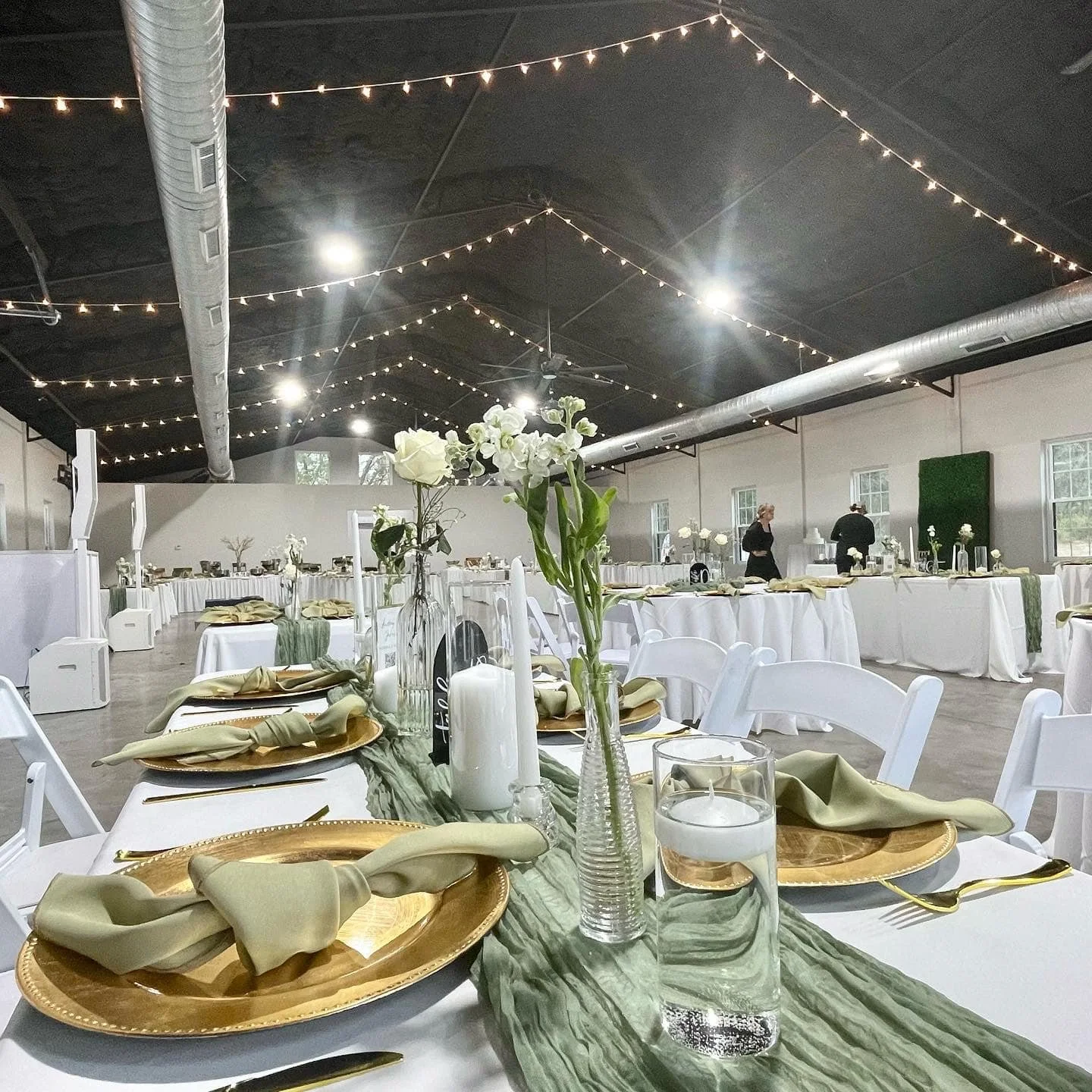 Elegant wedding reception setup with tables set with gold plates, green napkins, floral centerpieces, and candles under string lights.