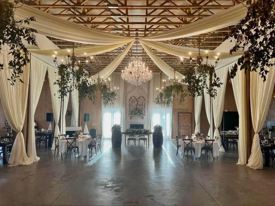 Elegant venue with draped curtains, chandeliers, round tables, chairs, and floral decorations.