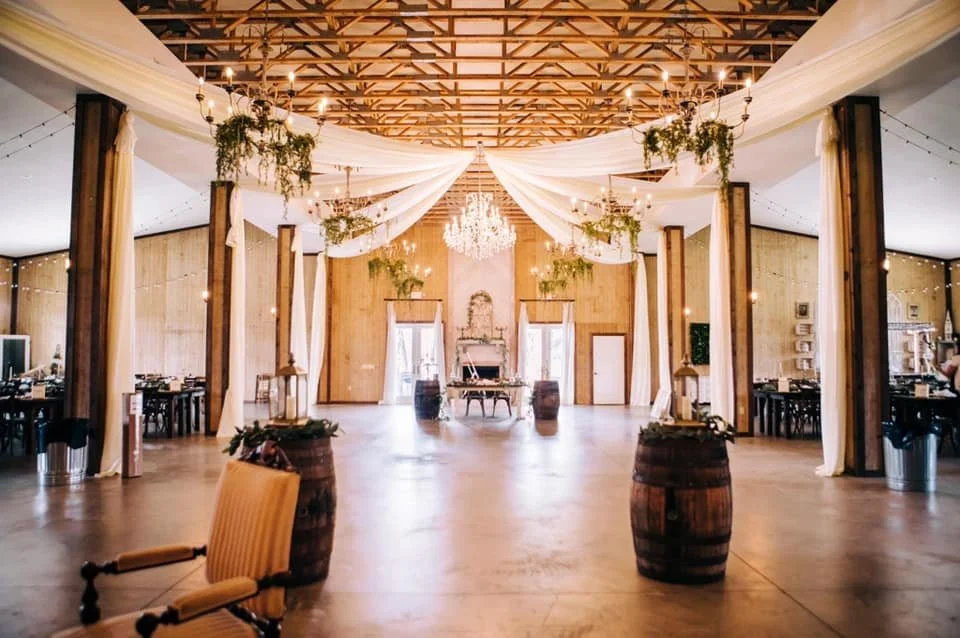 Rustic indoor wedding venue with wooden beams, draped fabric, chandeliers, and decorative barrels.