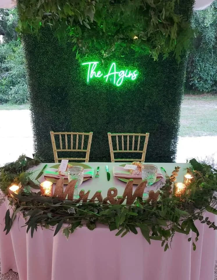 Wedding sweetheart table with green foliage, "The Agus" neon sign, and "Mr. & Mrs." wooden sign.